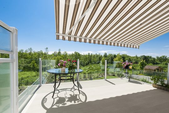 Tan striped retractable awning extended over a glass-railed balcony with a bistro table